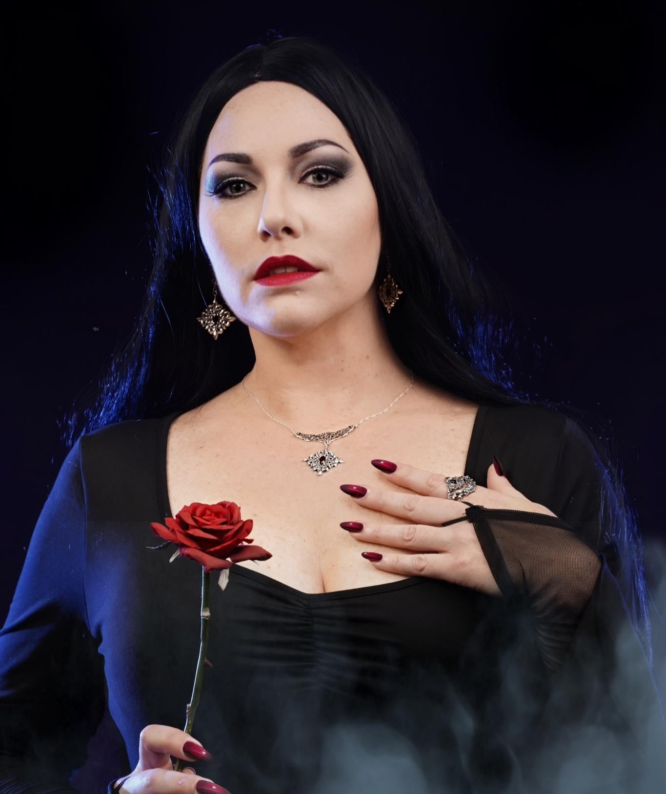 Woman with dark hair and red lipstick holding a red rose against a dark background. Morticia Addams portrayed by TooniBug.